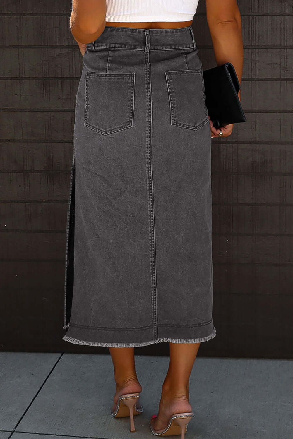 Back view of a black midi denim skirt with raw edge, side slits, and metal buttons, paired with a white top.