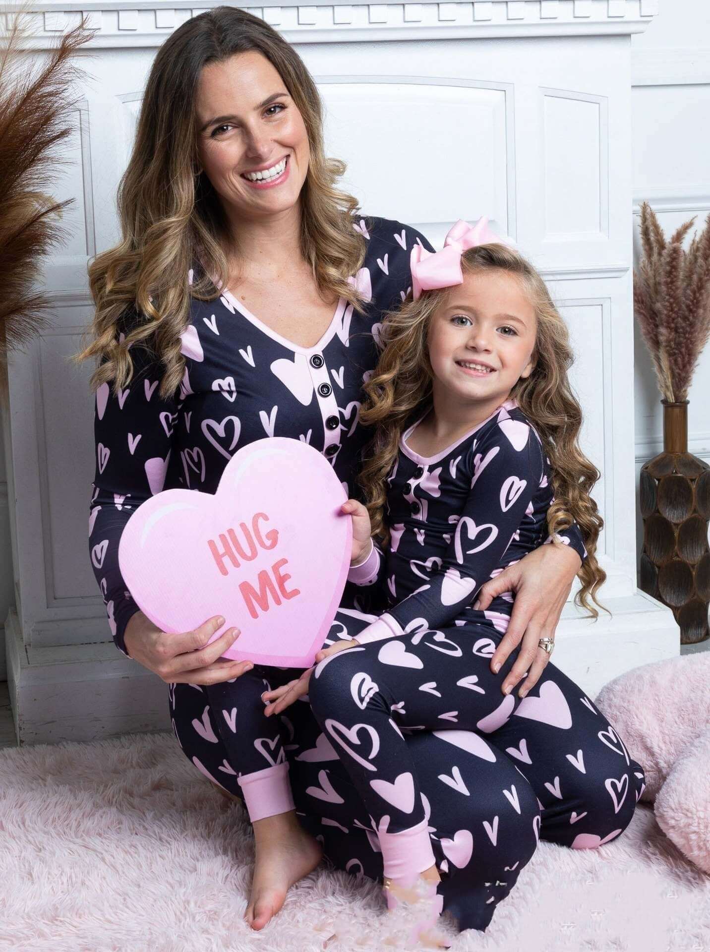 Mother and daughter in heart print pajamas holding a pink heart pillow that says 'HUG ME'.