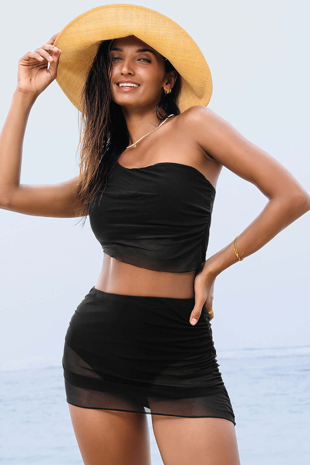 Black ruched mesh one shoulder bikini top and skirt set with a stylish sun hat at the beach.