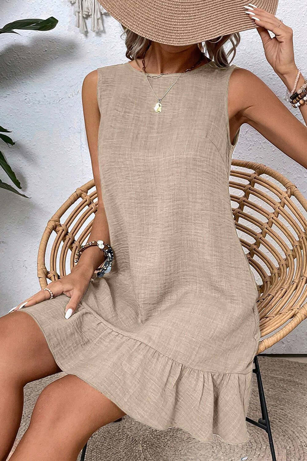 Model wearing a beige sleeveless mini dress with a ruffle hem, seated in a chair and holding a wide-brimmed hat.