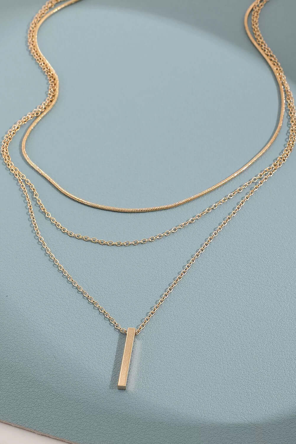 Gold bar pendant layered necklace with adjustable chains on a blue background.