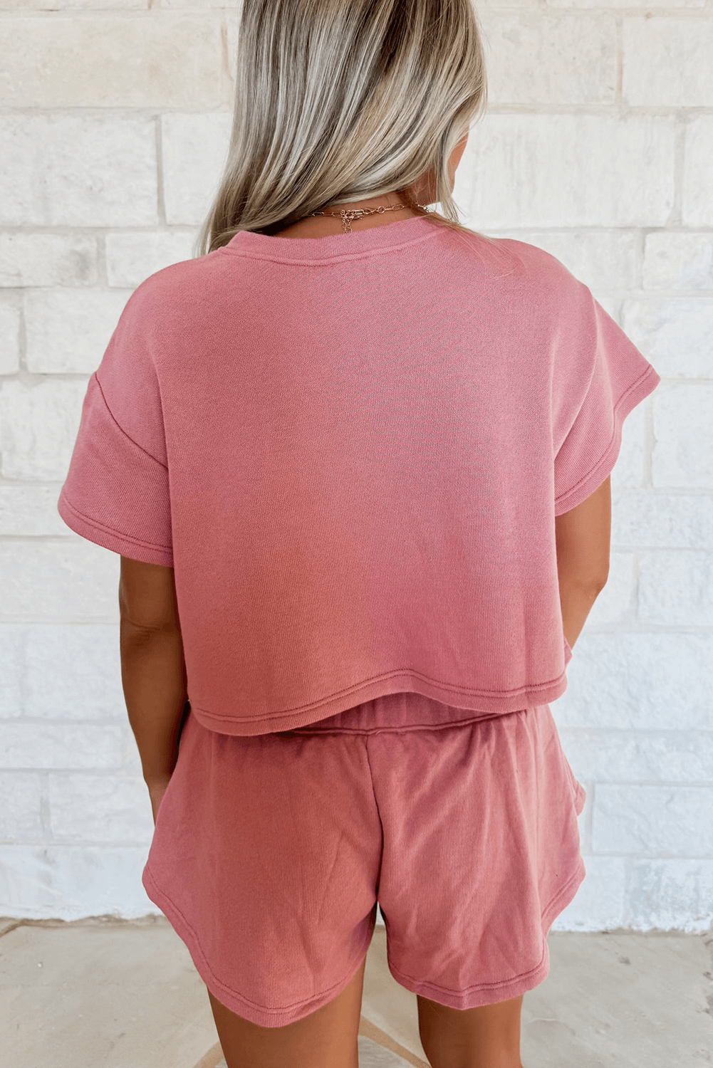 Back view of a girl wearing a pink cropped tee and matching ruffle high waist skort set against a textured wall.